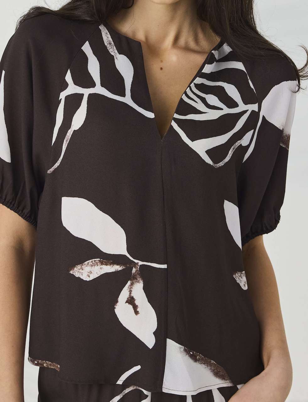 Lania The Label Model modeling a short-sleeve button-front blouse with a dark brown base and large white botanical leaf print, paired with matching dark pants, against a light gray studio backdrop with soft leaf shadows.