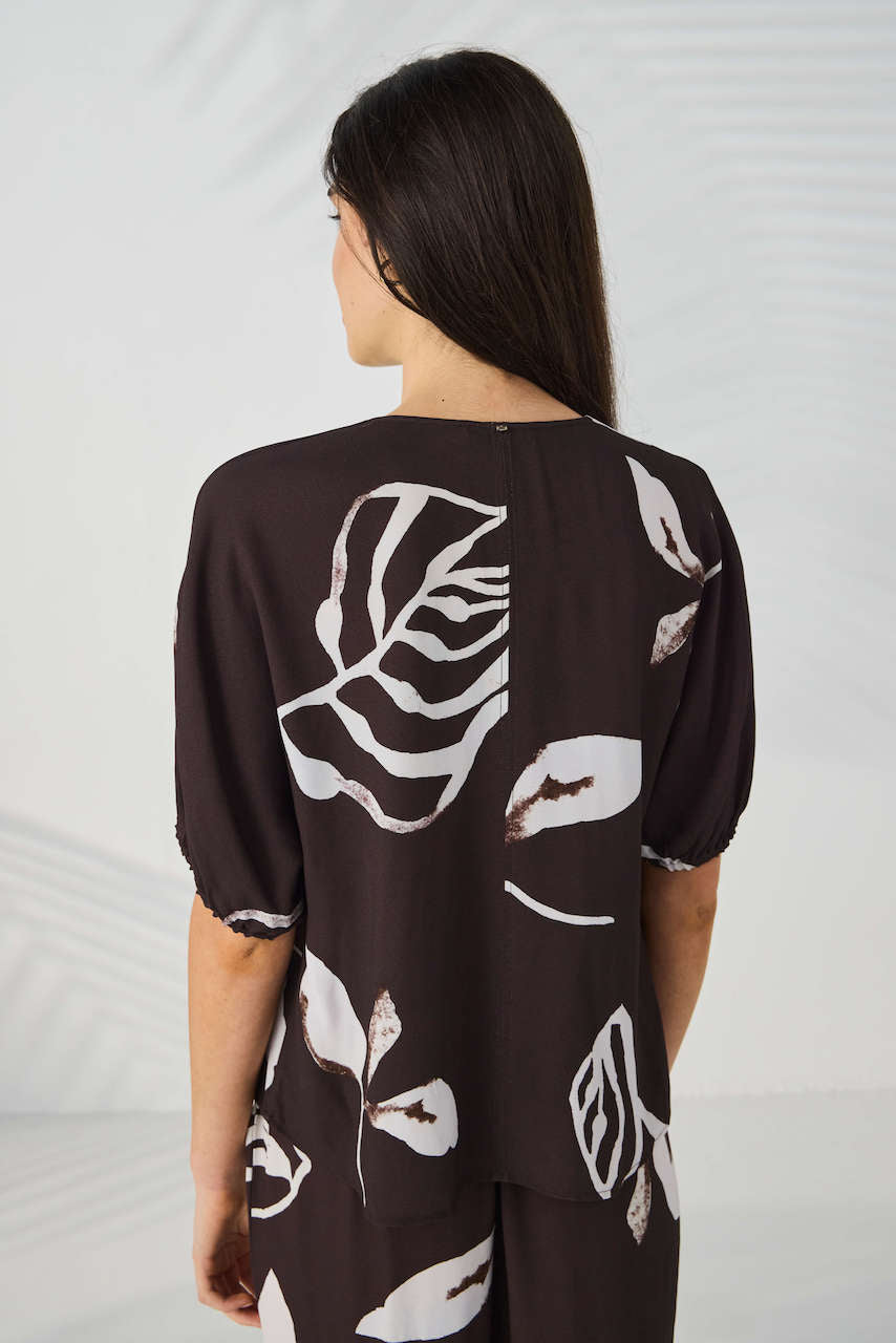 Lania The Label Model modeling a short-sleeve button-front blouse with a dark brown base and large white botanical leaf print, paired with matching dark pants, against a light gray studio backdrop with soft leaf shadows.