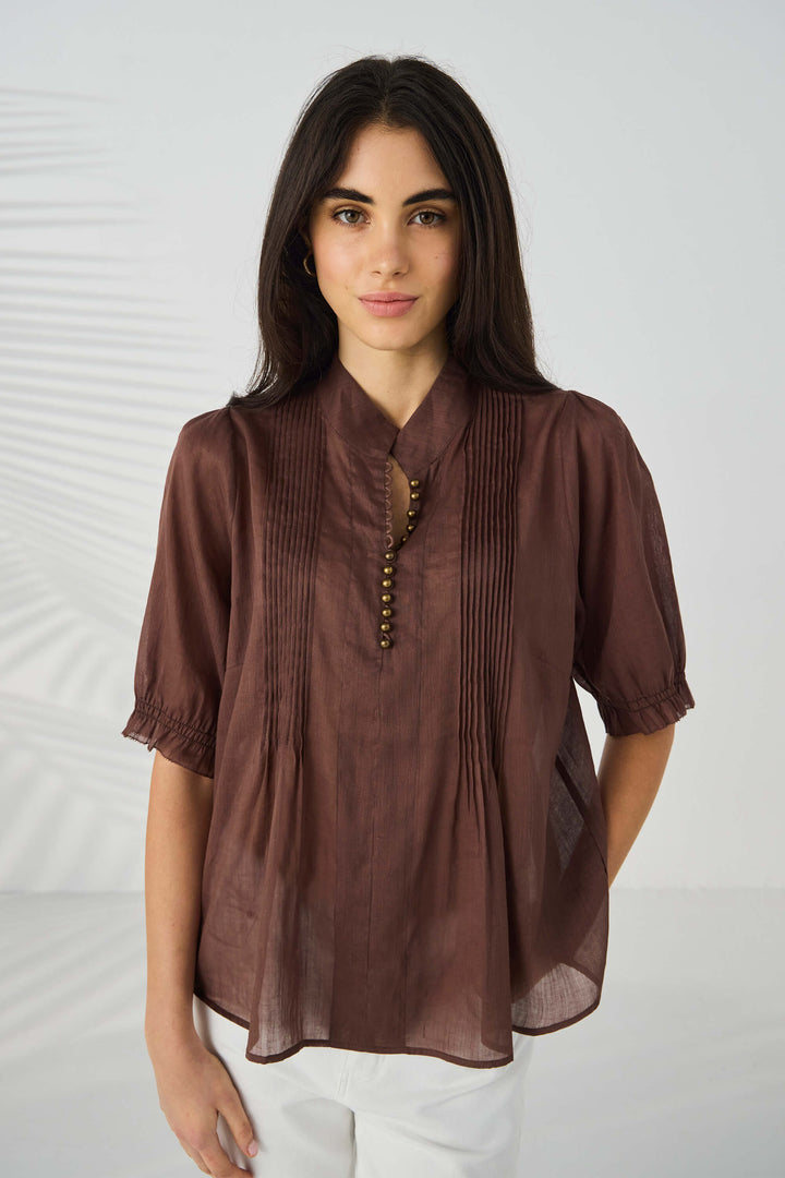 Lania The Label model wearing a brown short-sleeve blouse with pleated front detailing and a decorative beaded neckline, paired with white pants, against a light gray background with soft leaf shadows.