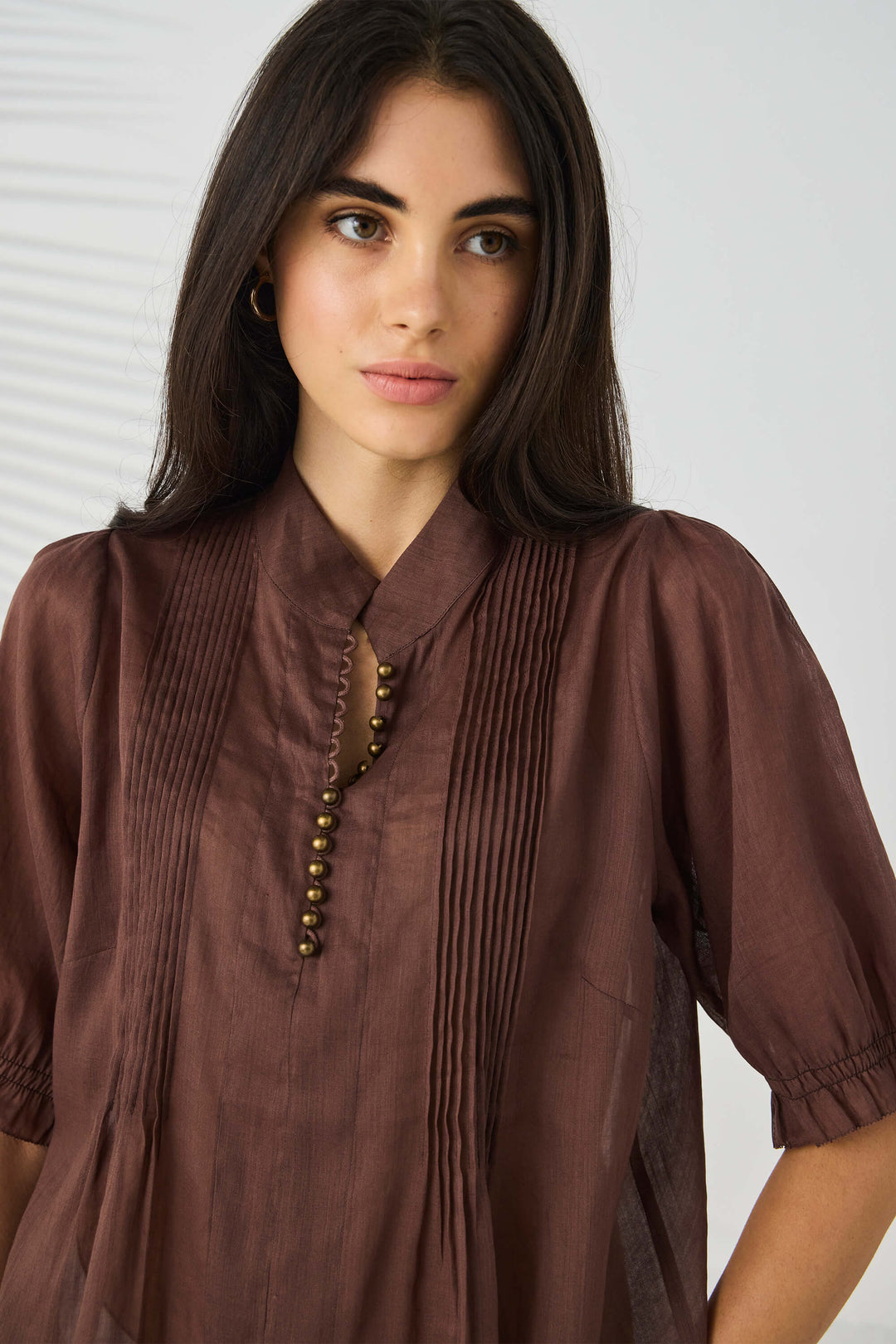 Lania The Label model wearing a brown short-sleeve blouse with pleated front detailing and a decorative beaded neckline, paired with white pants, against a light gray background with soft leaf shadows.