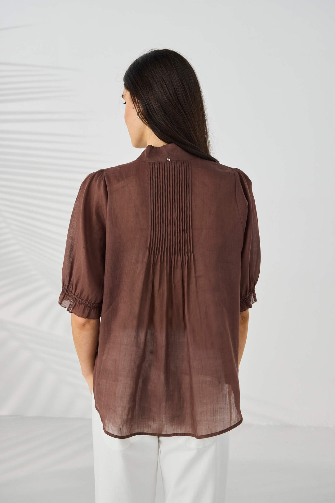 Lania The Label model wearing a brown short-sleeve blouse with pleated front detailing and a decorative beaded neckline, paired with white pants, against a light gray background with soft leaf shadows. Back view.