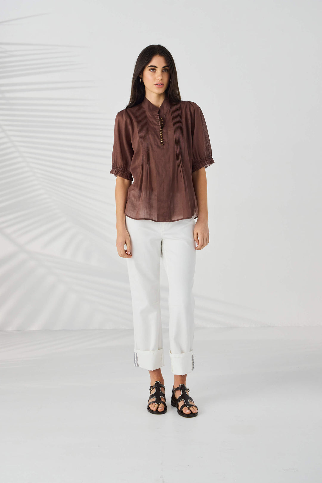 Lania The Label model wearing a brown short-sleeve blouse with pleated front detailing and a decorative beaded neckline, paired with white pants, against a light gray background with soft leaf shadows.