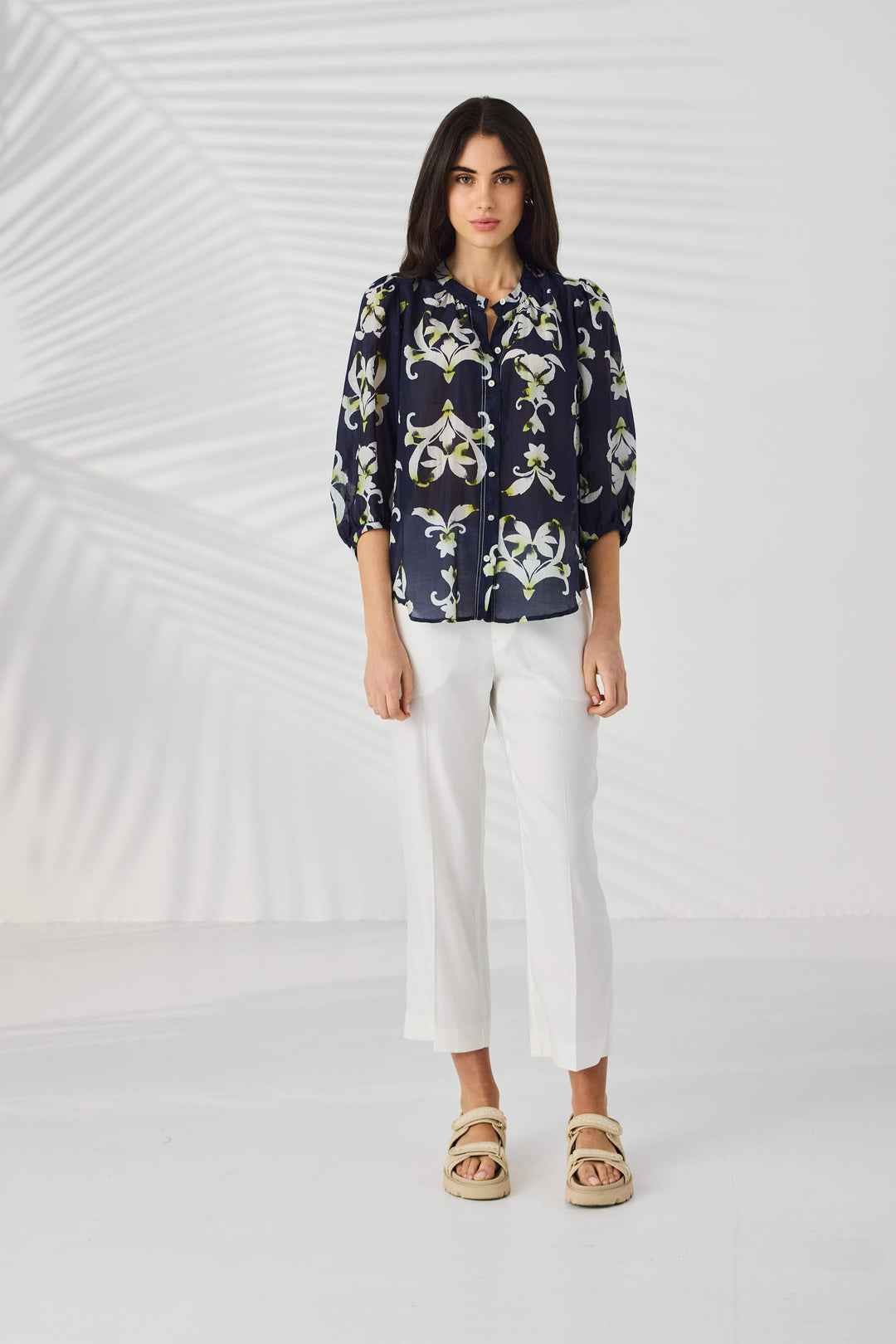 Lania The Label  modeling a navy blue button-up blouse with a white and light green floral/lily pattern, paired with white trousers, against a light gray studio background with subtle leaf-like shadows.