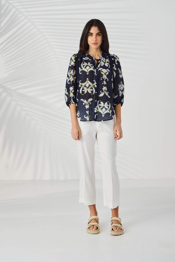 Lania The Label  modeling a navy blue button-up blouse with a white and light green floral/lily pattern, paired with white trousers, against a light gray studio background with subtle leaf-like shadows.