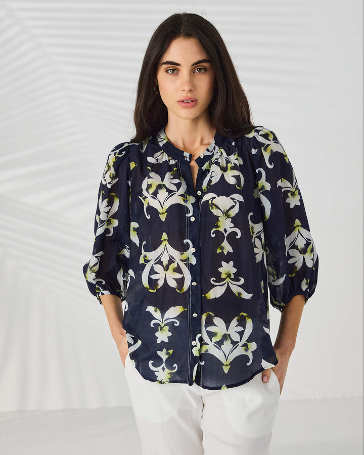 Lania The Label  modeling a navy blue button-up blouse with a white and light green floral/lily pattern, paired with white trousers, against a light gray studio background with subtle leaf-like shadows.