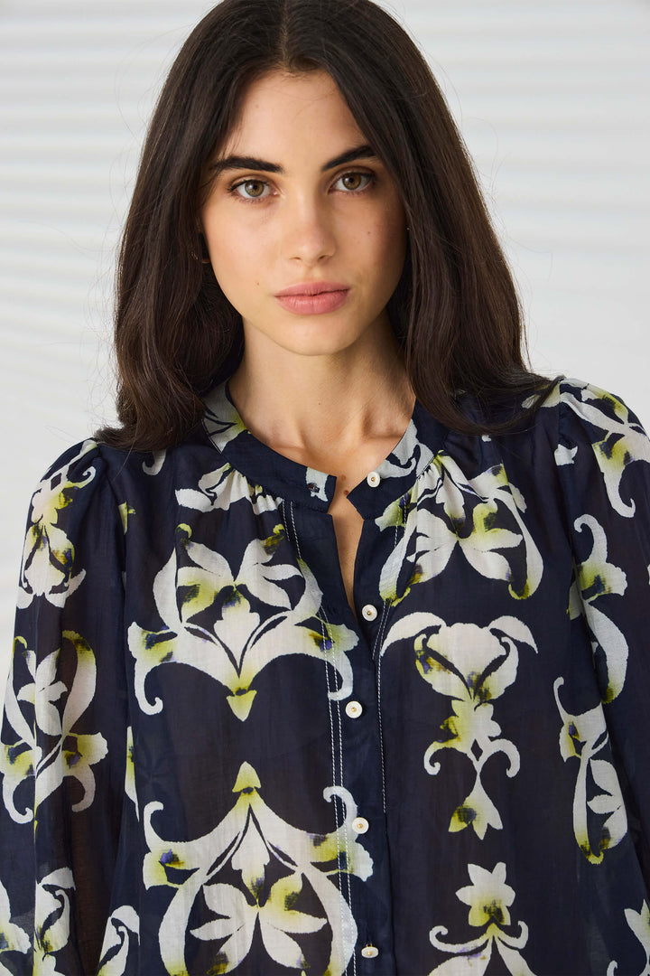 Lania The Label  modeling a navy blue button-up blouse with a white and light green floral/lily pattern, paired with white trousers, against a light gray studio background with subtle leaf-like shadows.