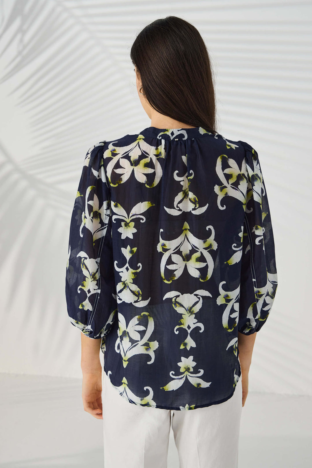 Lania The Label  modeling a navy blue button-up blouse with a white and light green floral/lily pattern, paired with white trousers, against a light gray studio background with subtle leaf-like shadows. Back view.