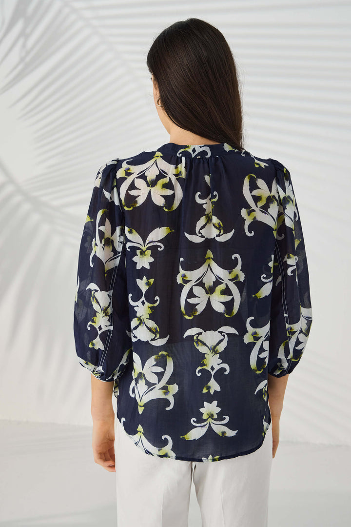 Lania The Label  modeling a navy blue button-up blouse with a white and light green floral/lily pattern, paired with white trousers, against a light gray studio background with subtle leaf-like shadows. Back view.
