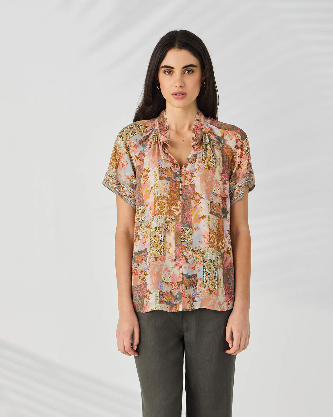 Lania The Label modeling a short-sleeve button-up blouse with a multicolor abstract and floral print in pink, beige, brown, and green tones, set against a light gray studio background.