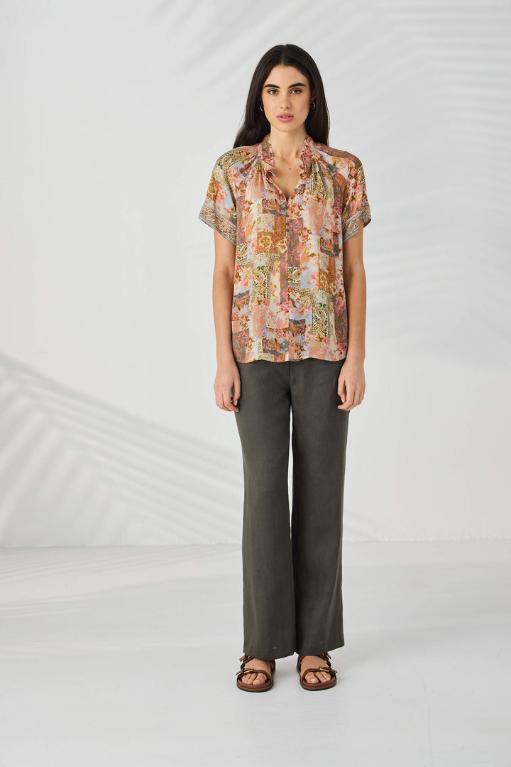 Lania The Label modeling a short-sleeve button-up blouse with a multicolor abstract and floral print in pink, beige, brown, and green tones, set against a light gray studio background.