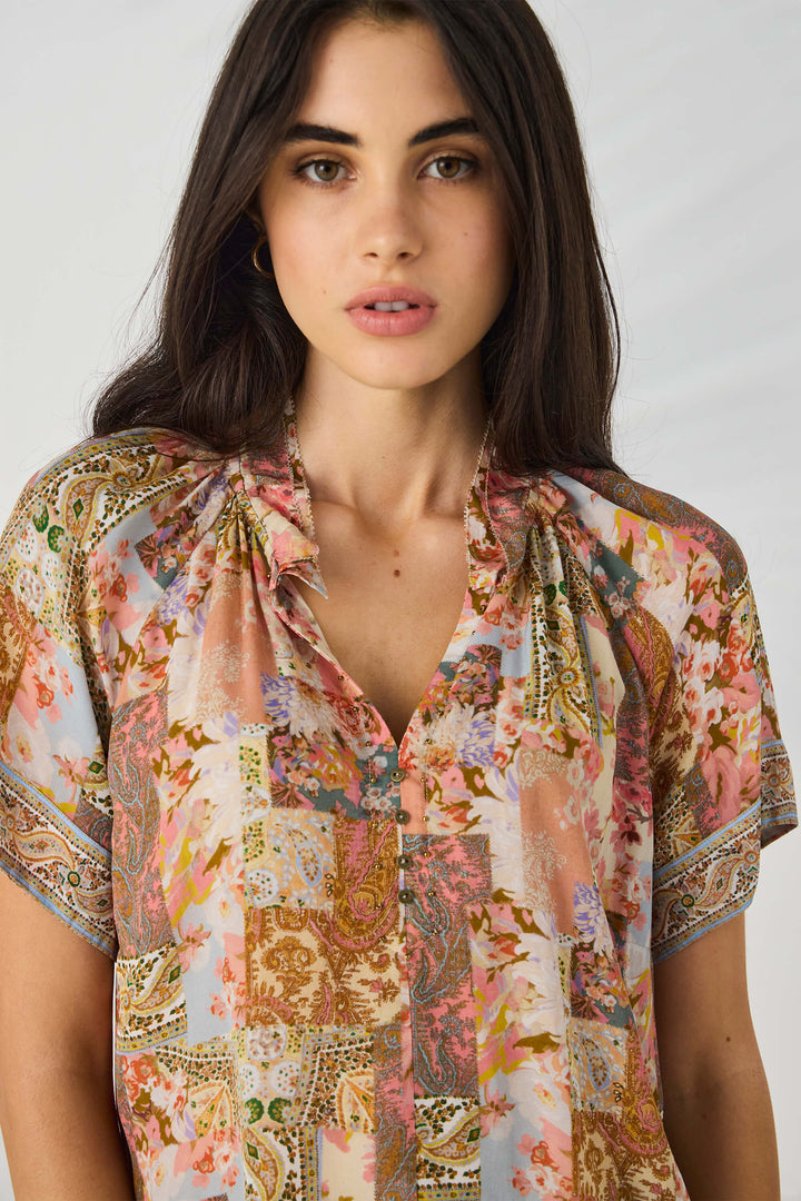Lania The Label modeling a short-sleeve button-up blouse with a multicolor abstract and floral print in pink, beige, brown, and green tones, set against a light gray studio background.