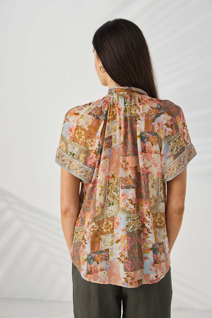 Lania The Label modeling a short-sleeve button-up blouse with a multicolor abstract and floral print in pink, beige, brown, and green tones, set against a light gray studio background. Back view.