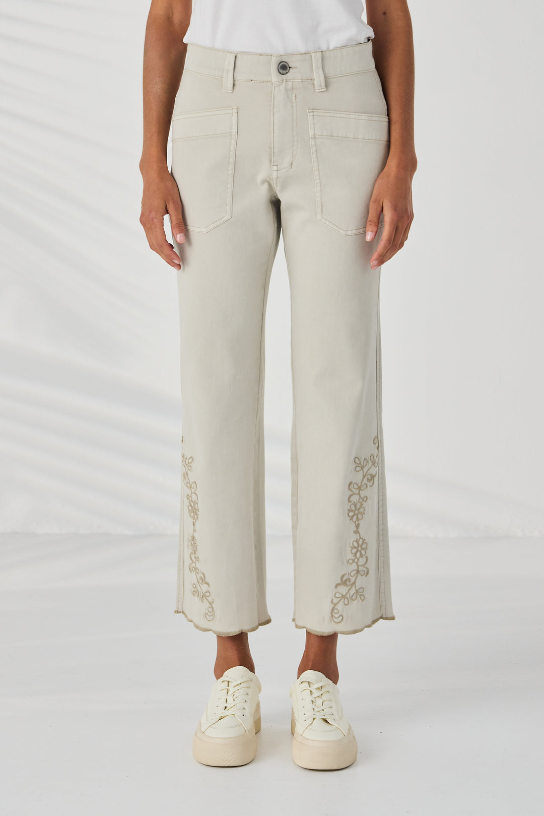 Lania The Label A model is wearing a pair of light beige cropped jeans with gold floral embroidery on the lower legs, paired with a white t-shirt and cream platform sneakers against a white studio background.