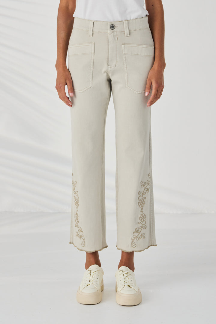 Lania The Label A model is wearing a pair of light beige cropped jeans with gold floral embroidery on the lower legs, paired with a white t-shirt and cream platform sneakers against a white studio background.
