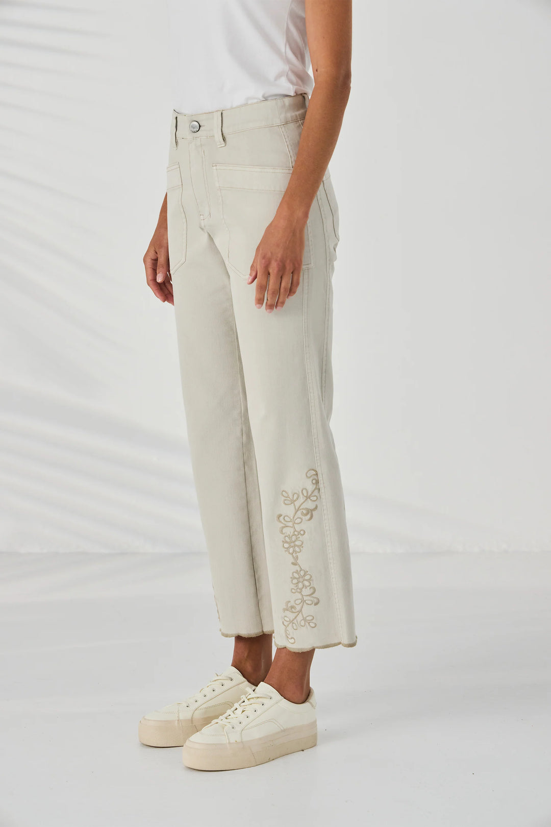 Lania The Label A model is wearing a pair of light beige cropped jeans with gold floral embroidery on the lower legs, paired with a white t-shirt and cream platform sneakers against a white studio background.