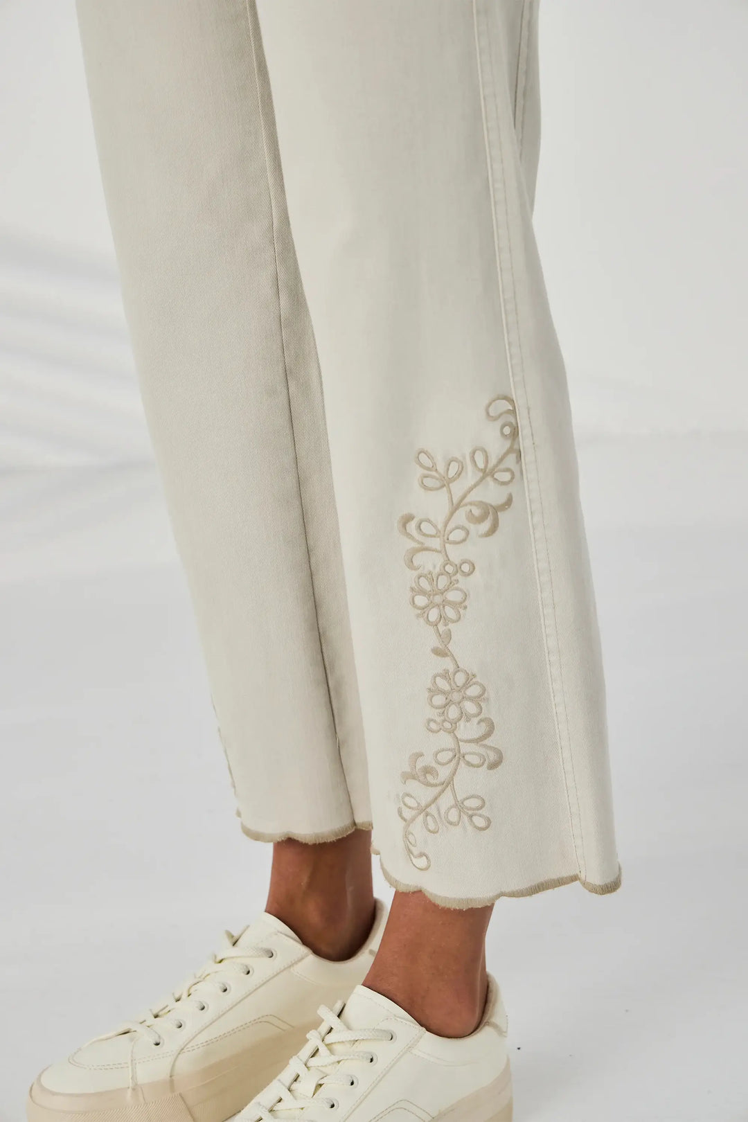 Lania The Label A model is wearing a pair of light beige cropped jeans with gold floral embroidery on the lower legs, paired with a white t-shirt and cream platform sneakers against a white studio background.