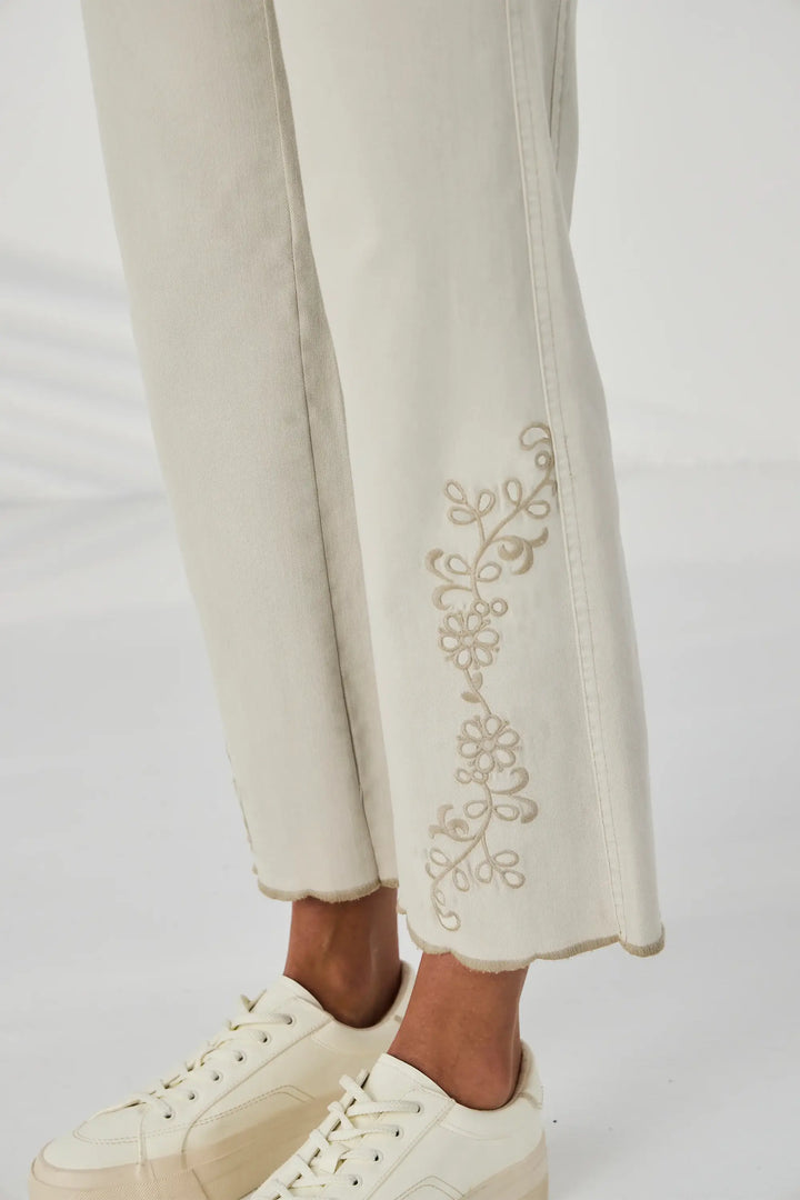 Lania The Label A model is wearing a pair of light beige cropped jeans with gold floral embroidery on the lower legs, paired with a white t-shirt and cream platform sneakers against a white studio background.