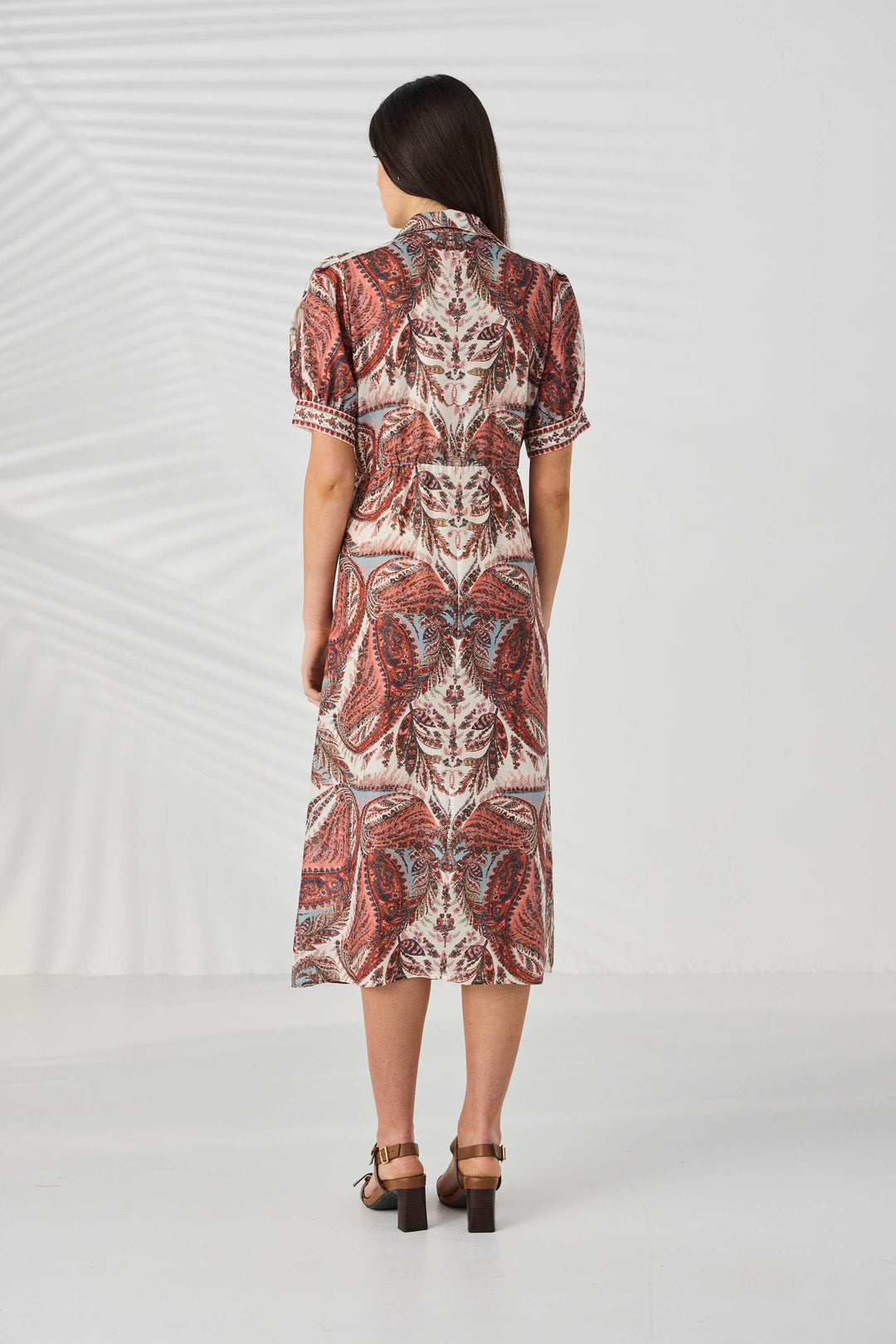 Lania The Label model viewed from behind wearing a short-sleeve midi dress with a detailed red, brown, and beige paisley pattern, paired with brown block-heel sandals, set against a white studio background with subtle leaf shadows.