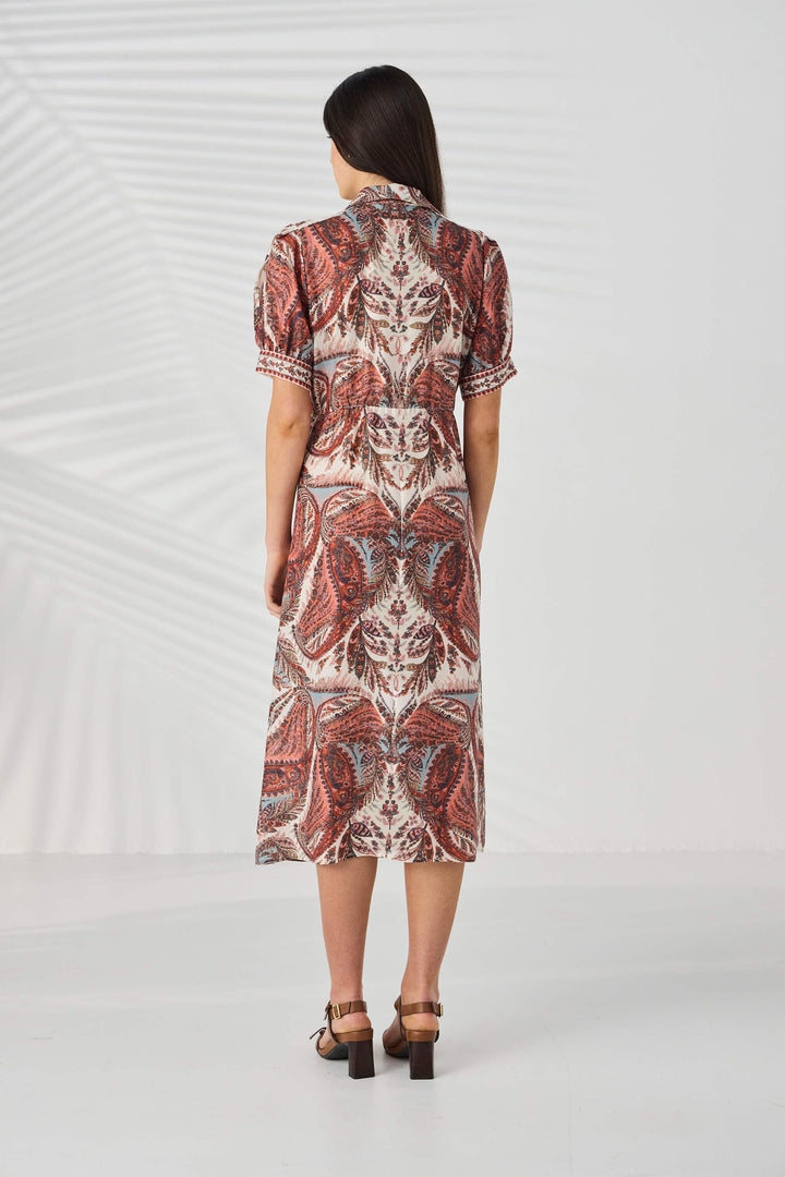 Lania The Label model viewed from behind wearing a short-sleeve midi dress with a detailed red, brown, and beige paisley pattern, paired with brown block-heel sandals, set against a white studio background with subtle leaf shadows.