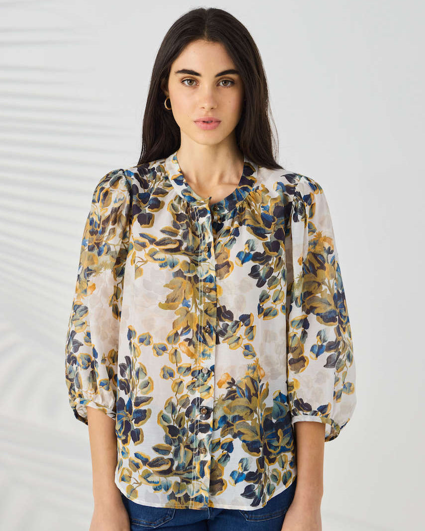 Lania The Label modeling a lightweight floral blouse with a cream base and blue, olive green, and mustard yellow botanical print, paired with blue jeans against a light gray studio background.