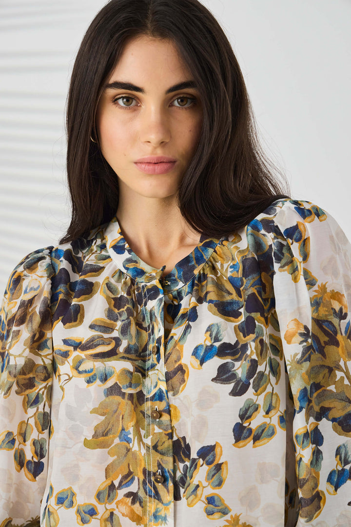 Lania The Label modeling a lightweight floral blouse with a cream base and blue, olive green, and mustard yellow botanical print, paired with blue jeans against a light gray studio background.