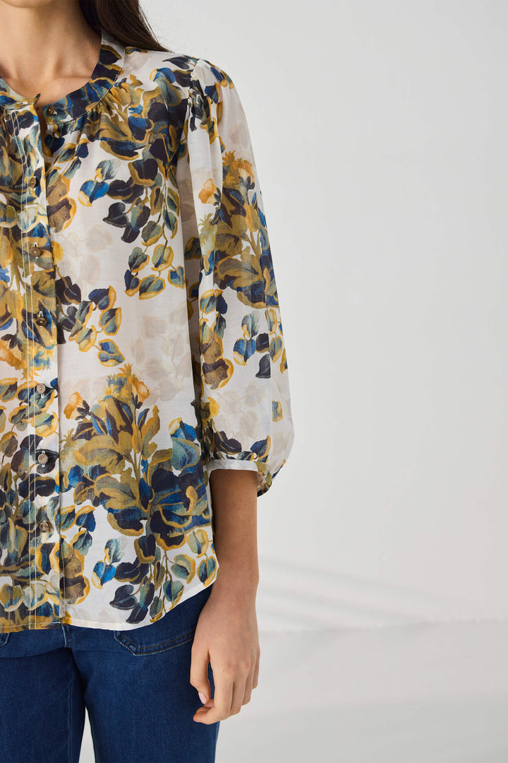Lania The Label modeling a lightweight floral blouse with a cream base and blue, olive green, and mustard yellow botanical print, paired with blue jeans against a light gray studio background.