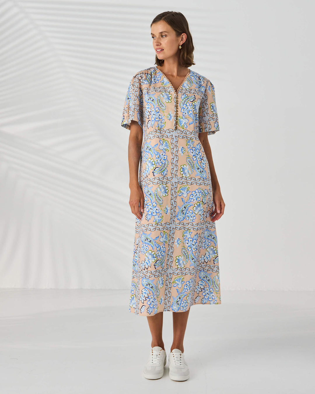 Lania The label model in a short-sleeve midi dress with a blue and beige paisley pattern, standing against a white studio background with leaf shadows.