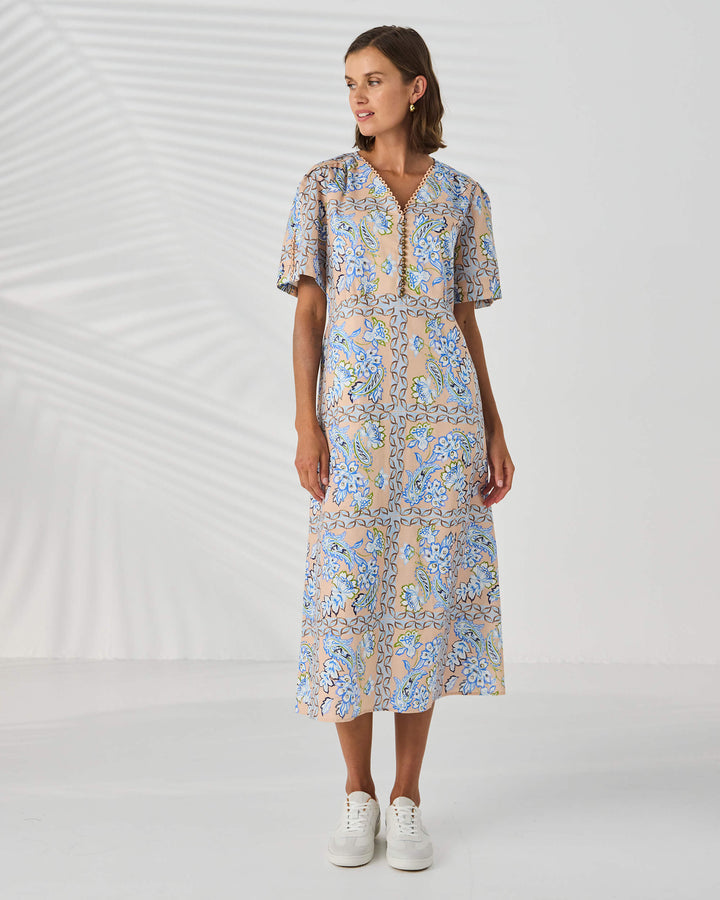 Lania The label model in a short-sleeve midi dress with a blue and beige paisley pattern, standing against a white studio background with leaf shadows.