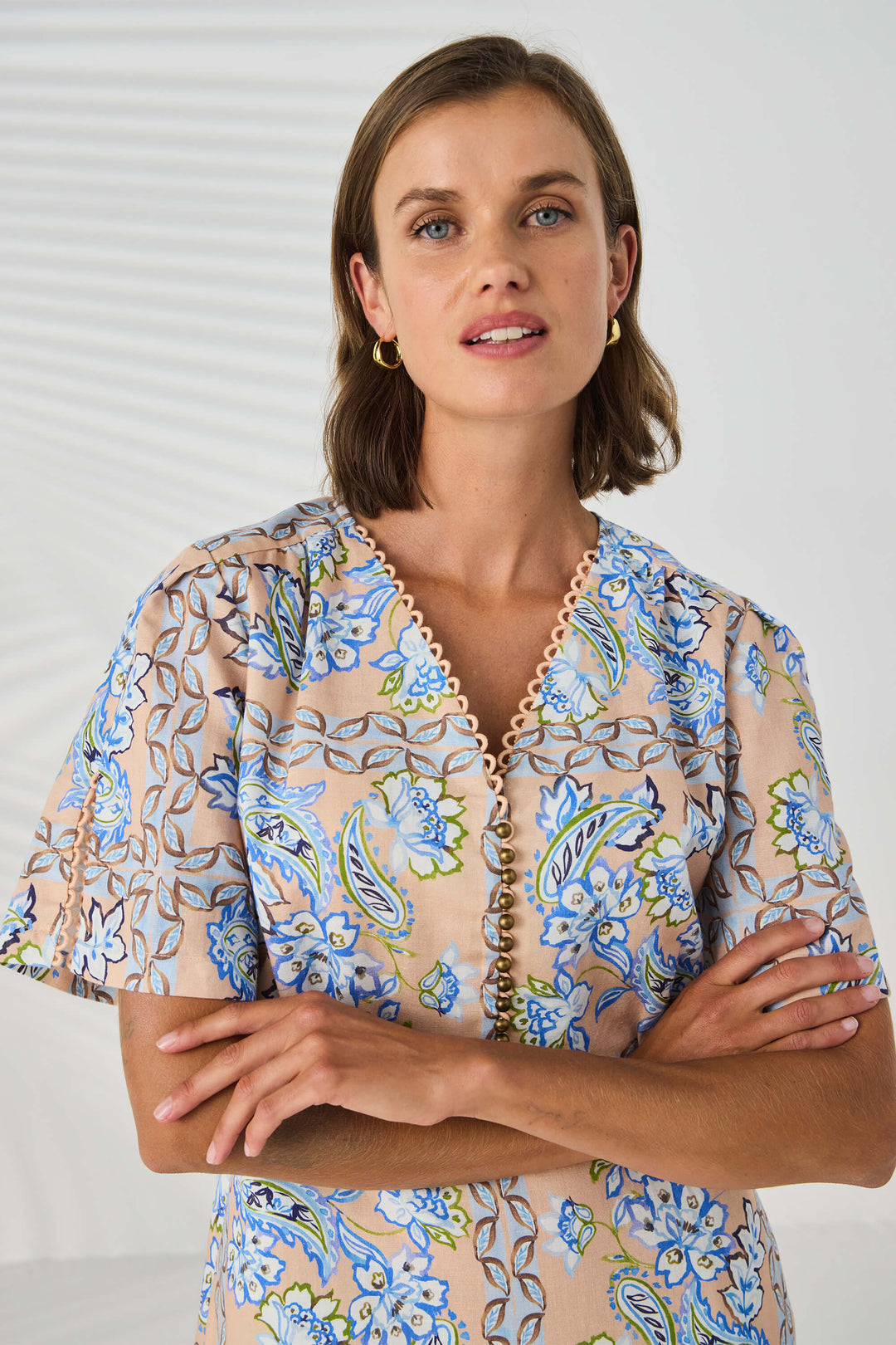 Lania The label model in a short-sleeve midi dress with a blue and beige paisley pattern, standing against a white studio background with leaf shadows.