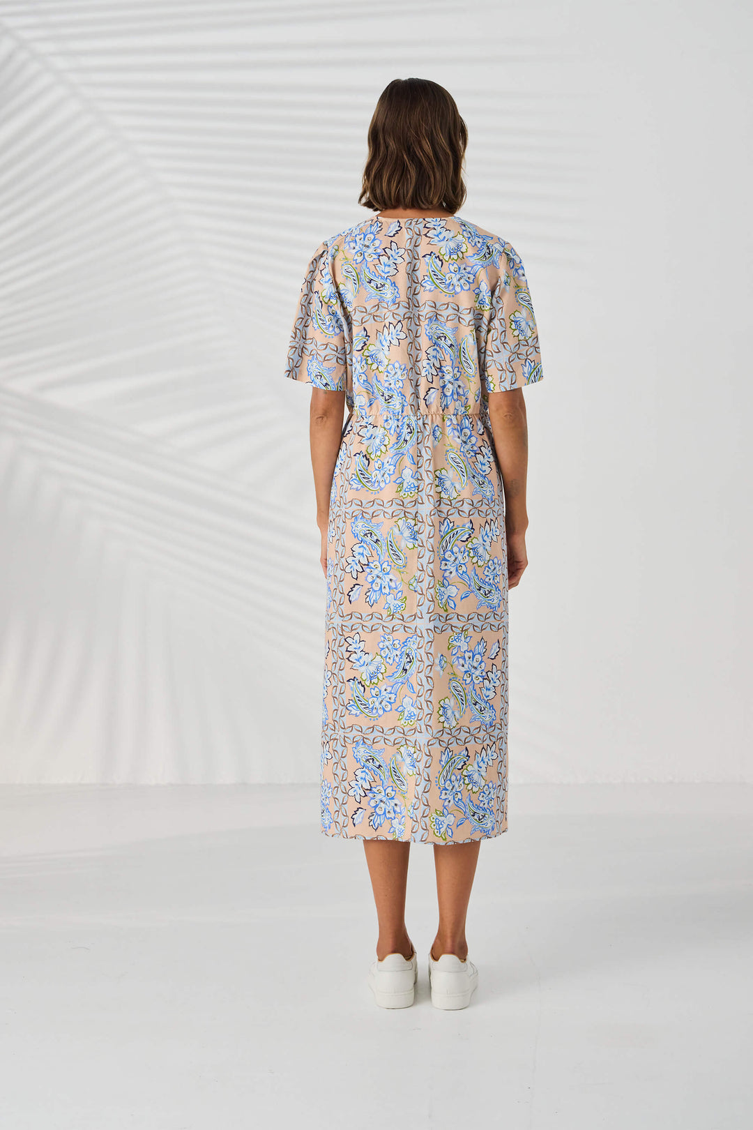 Lania The label model in a short-sleeve midi dress with a blue and beige paisley pattern, standing against a white studio background with leaf shadows. Back view.