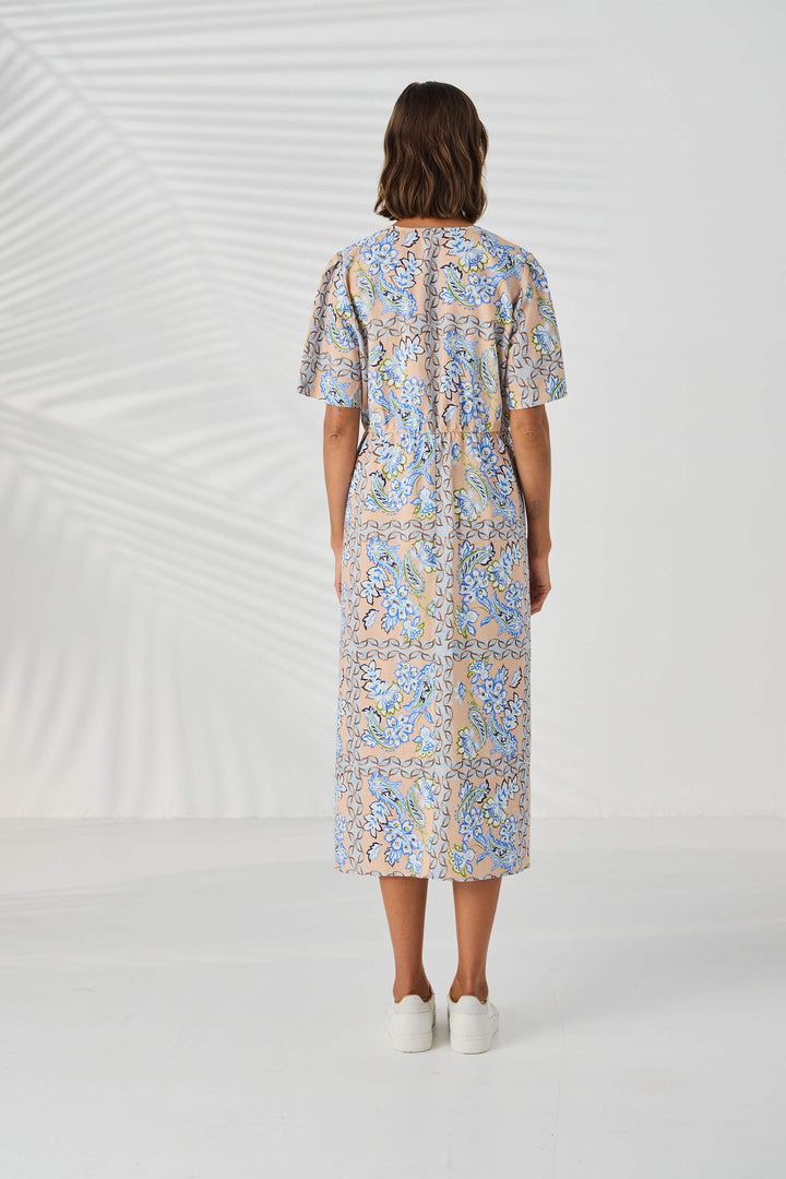 Lania The label model in a short-sleeve midi dress with a blue and beige paisley pattern, standing against a white studio background with leaf shadows. Back view.