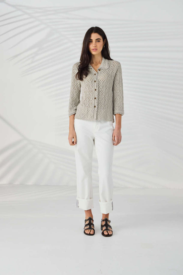 Lania The Label Model wearing a light beige open-knit button-up cardigan with white pants, standing against a white background with subtle leaf shadows.