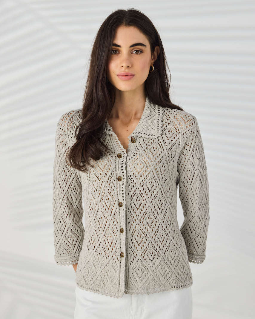 Lania The Label Model wearing a light beige open-knit button-up cardigan with white pants, standing against a white background with subtle leaf shadows.