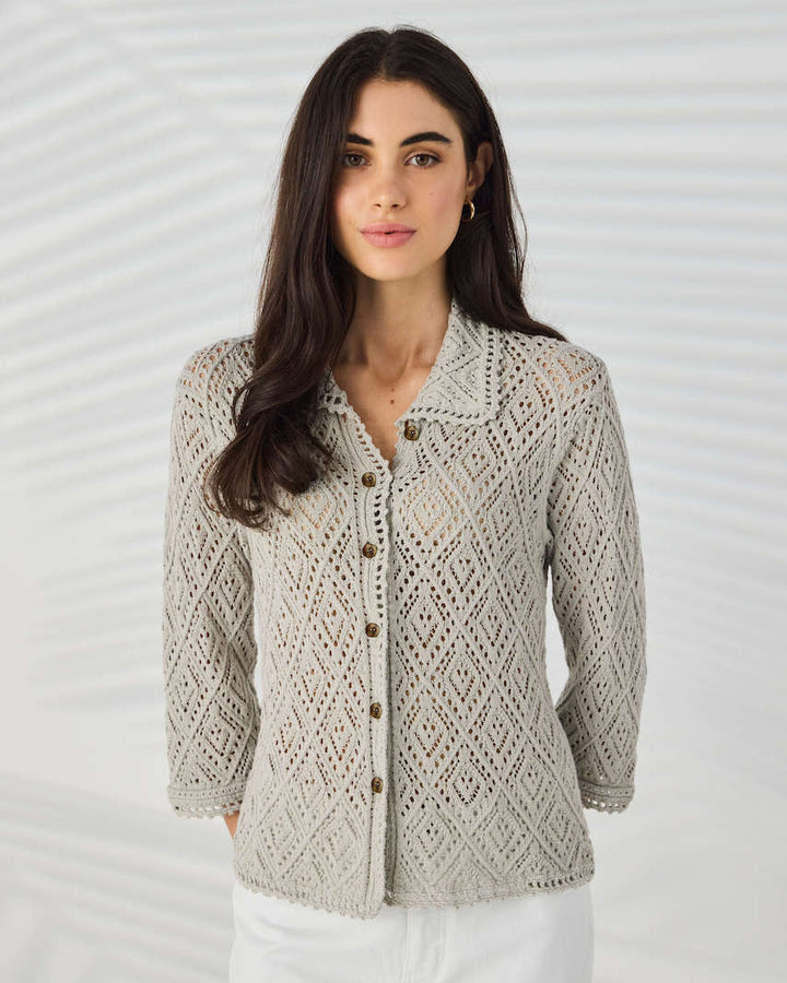 Lania The Label Model wearing a light beige open-knit button-up cardigan with white pants, standing against a white background with subtle leaf shadows.