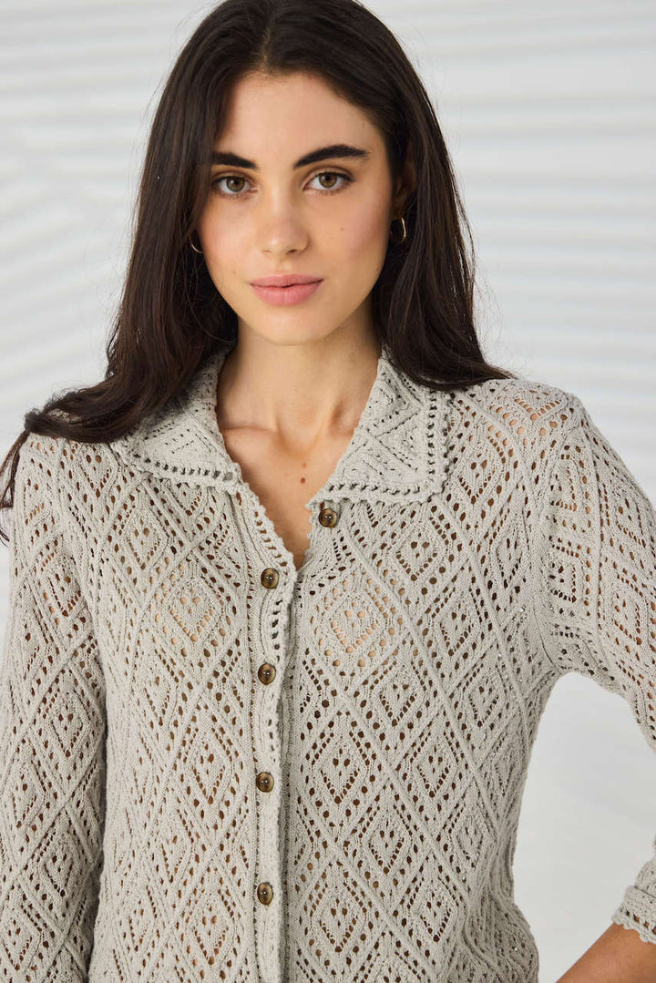Lania The Label Model wearing a light beige open-knit button-up cardigan with white pants, standing against a white background with subtle leaf shadows.