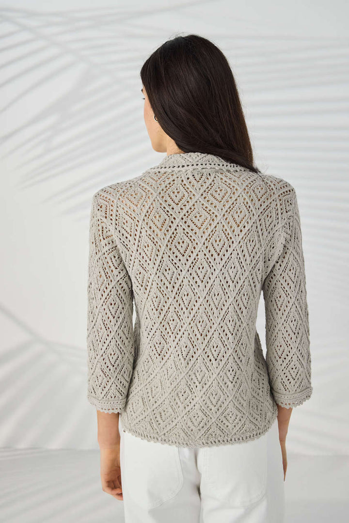 Lania The Label Model wearing a light beige open-knit button-up cardigan with white pants, standing against a white background with subtle leaf shadows.