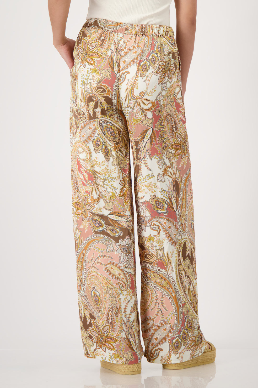 Monari model is shown from the waist down wearing wide-leg trousers with a detailed paisley pattern in shades of beige, brown, pink, and gold. The pants feature an elastic drawstring waist and side pockets, paired with platform sandals on a light gray studio background. Back view 