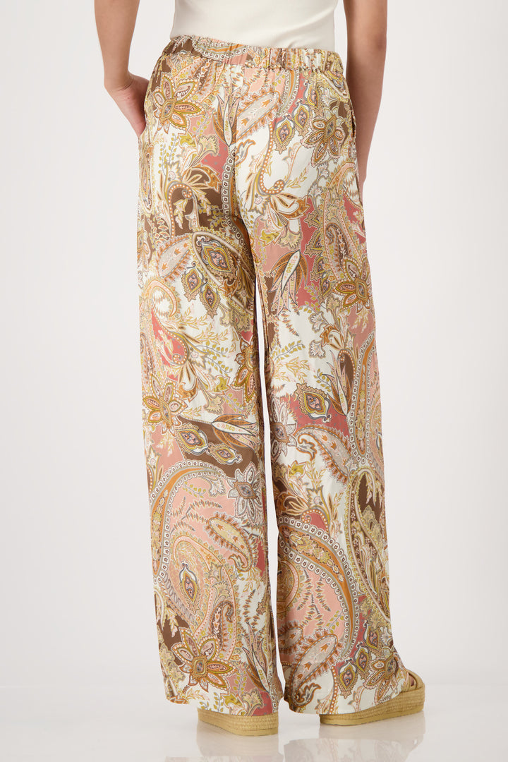 Monari model is shown from the waist down wearing wide-leg trousers with a detailed paisley pattern in shades of beige, brown, pink, and gold. The pants feature an elastic drawstring waist and side pockets, paired with platform sandals on a light gray studio background. Back view 