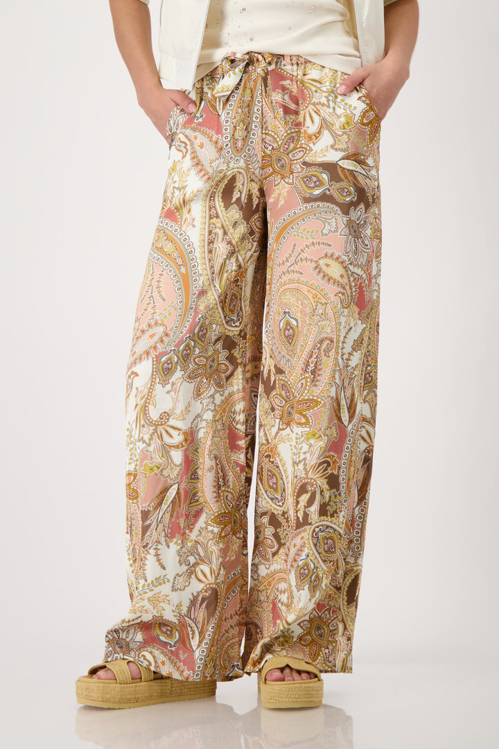 Monari model is shown from the waist down wearing wide-leg trousers with a detailed paisley pattern in shades of beige, brown, pink, and gold. The pants feature an elastic drawstring waist and side pockets, paired with platform sandals on a light gray studio background.