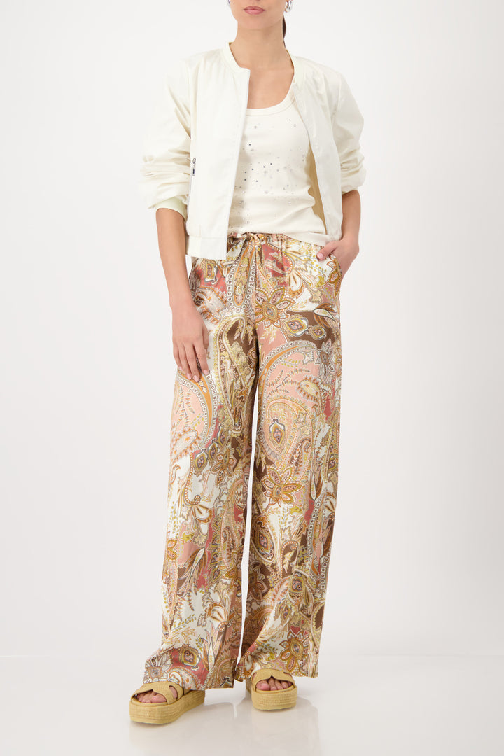 Monari model is shown from the waist down wearing wide-leg trousers with a detailed paisley pattern in shades of beige, brown, pink, and gold. The pants feature an elastic drawstring waist and side pockets, paired with platform sandals on a light gray studio background.