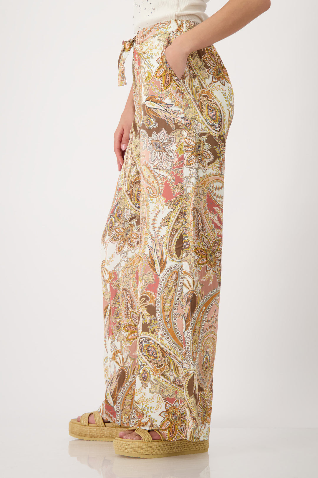 Monari model is shown from the waist down wearing wide-leg trousers with a detailed paisley pattern in shades of beige, brown, pink, and gold. The pants feature an elastic drawstring waist and side pockets, paired with platform sandals on a light gray studio background.