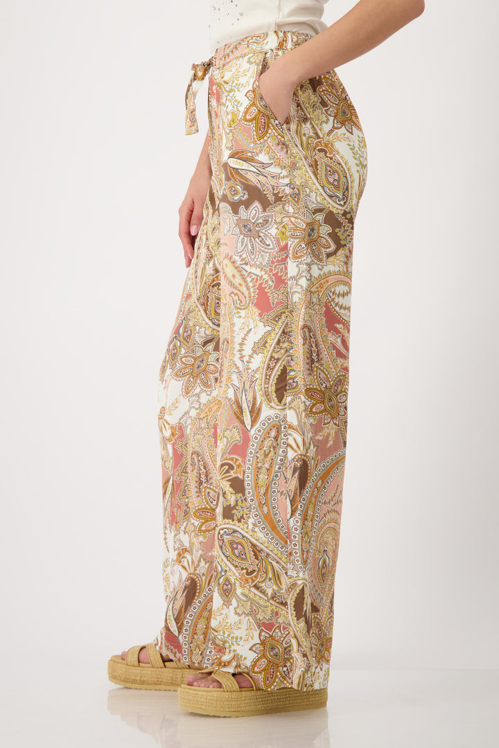 Monari model is shown from the waist down wearing wide-leg trousers with a detailed paisley pattern in shades of beige, brown, pink, and gold. The pants feature an elastic drawstring waist and side pockets, paired with platform sandals on a light gray studio background.