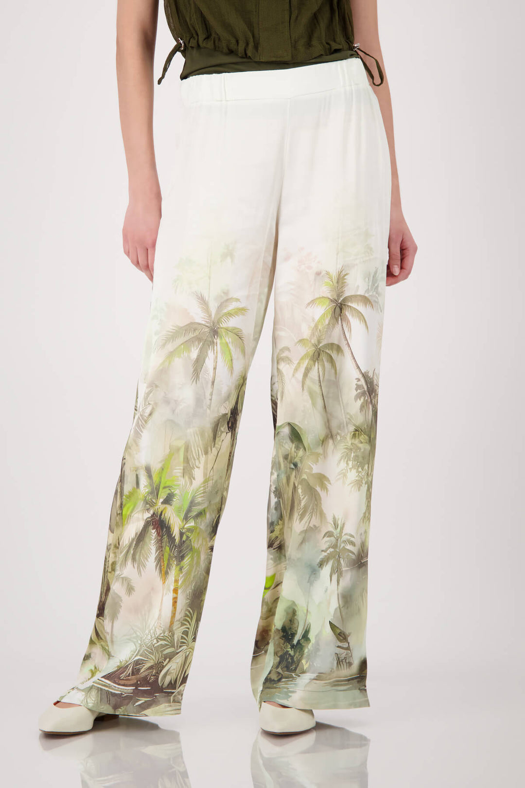 Monari woman modeling a short-sleeve olive green knit top paired with white wide-leg pants featuring a green palm tree print, set against a plain light gray studio background.