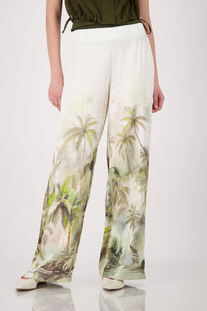 Monari woman modeling a short-sleeve olive green knit top paired with white wide-leg pants featuring a green palm tree print, set against a plain light gray studio background.