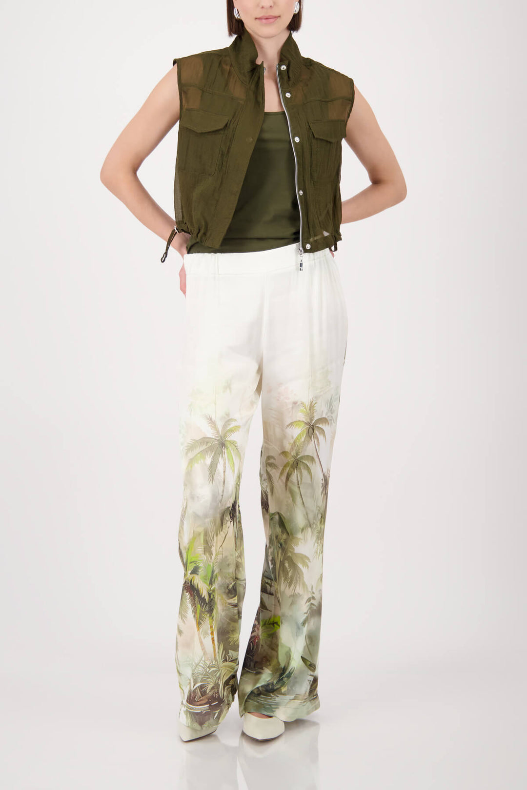 Monari woman modeling a short-sleeve olive green knit top paired with white wide-leg pants featuring a green palm tree print, set against a plain light gray studio background.