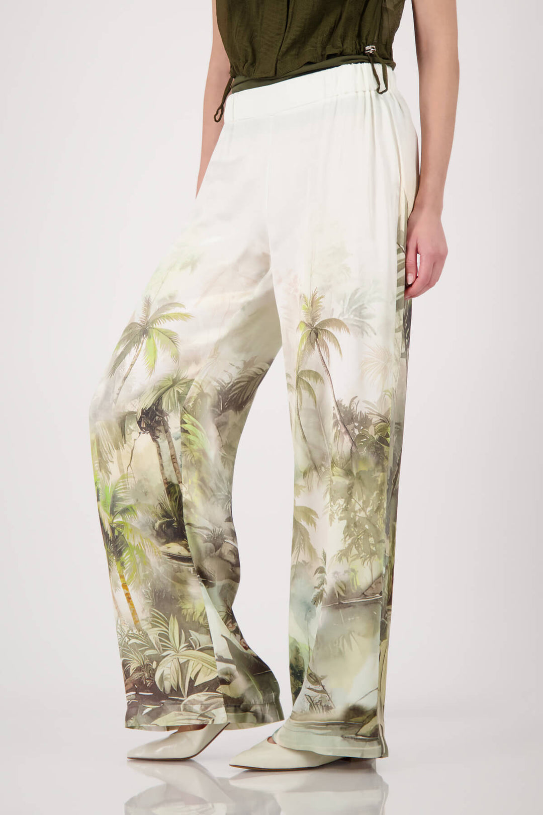 Monari woman modeling a short-sleeve olive green knit top paired with white wide-leg pants featuring a green palm tree print, set against a plain light gray studio background.