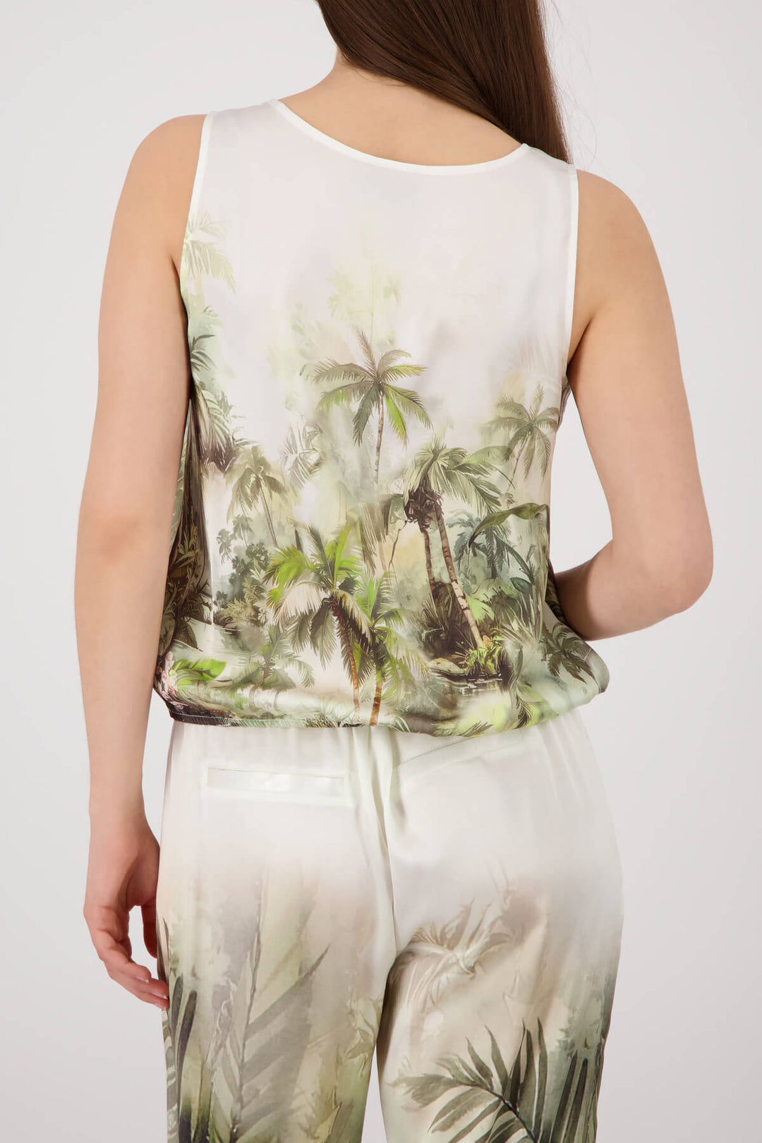Monari modeling a sleeveless white tank top with a green and brown palm tree tropical print, paired with matching white pants, against a light gray studio background. back view