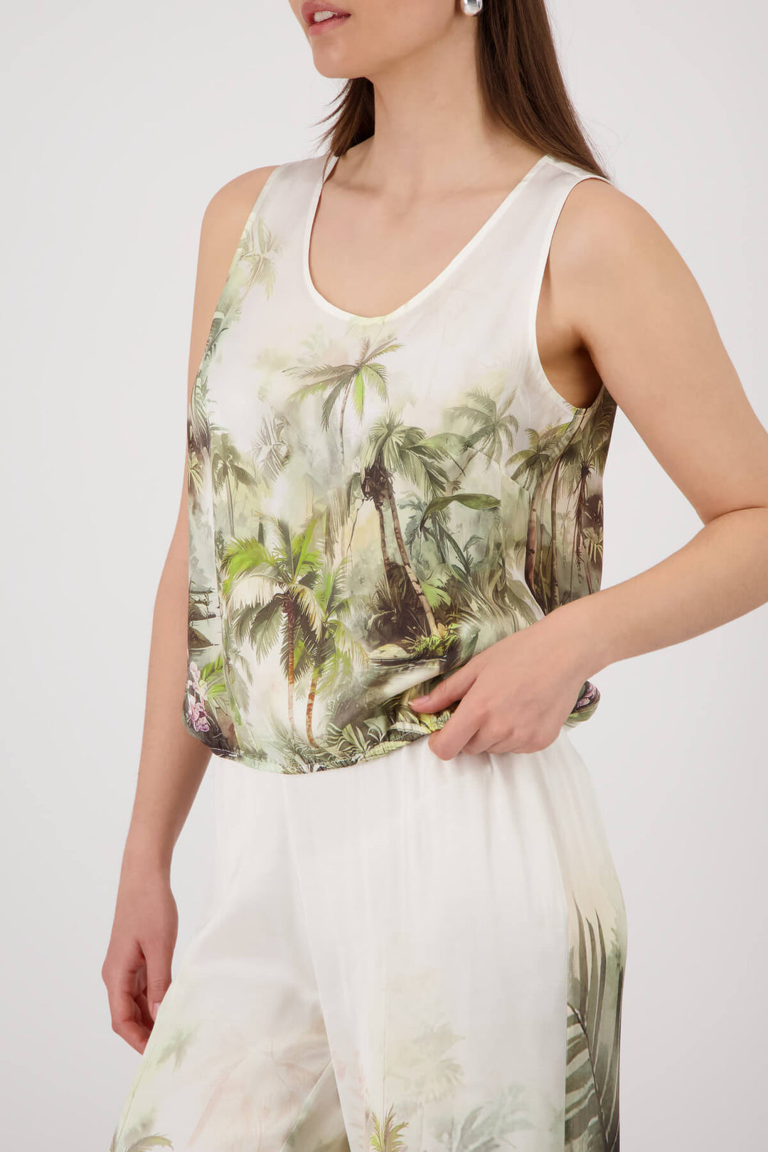 Monari modeling a sleeveless white tank top with a green and brown palm tree tropical print, paired with matching white pants, against a light gray studio background.