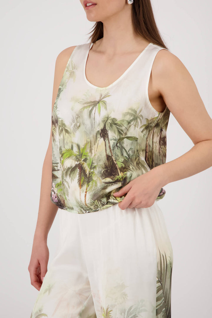 Monari modeling a sleeveless white tank top with a green and brown palm tree tropical print, paired with matching white pants, against a light gray studio background.