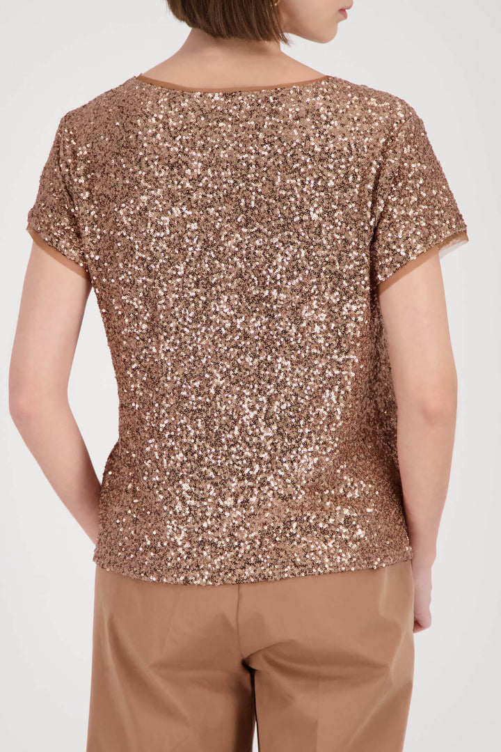 Monari modeling a short-sleeve crewneck top covered in shimmering rose gold sequins, paired with brown trousers, against a plain light gray studio background. Back view